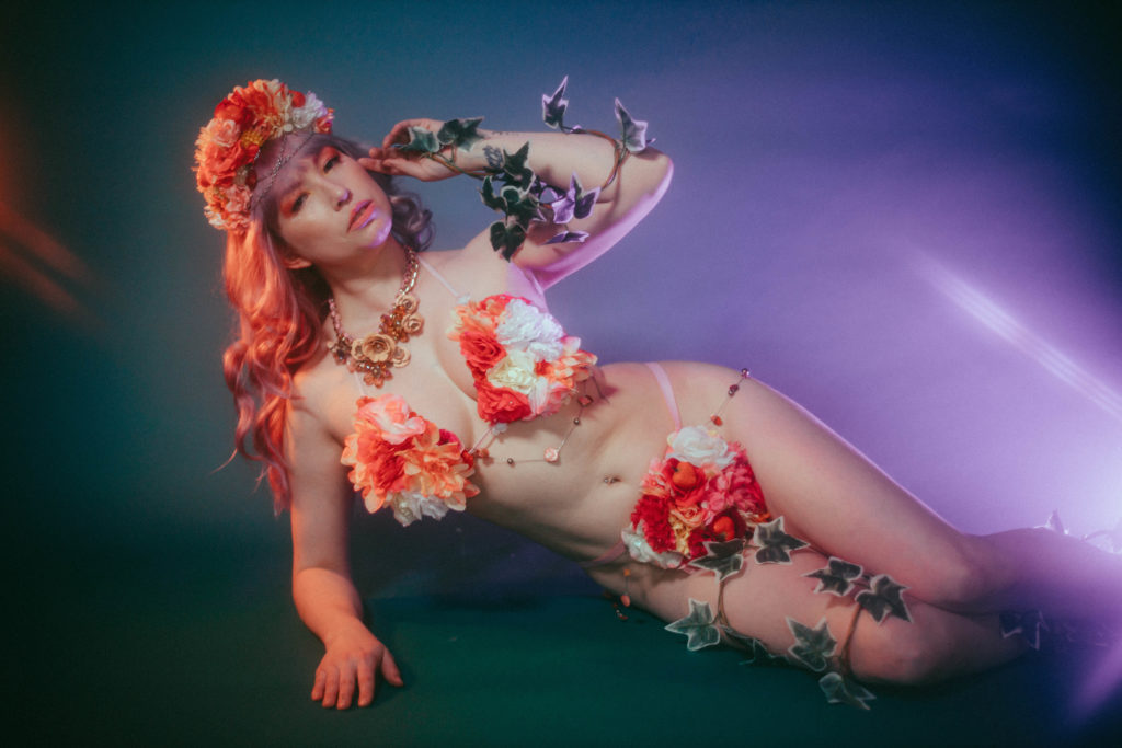 Color photo of Verronica Kirei laying on the floor wearing flower headdress, flower bra, and flower bottoms. 
