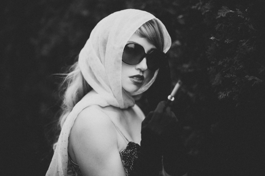 Black and white photo of Verronica Kirei wearing head scarf and sunglasses holding a cigarette.